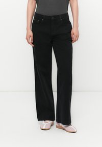Person iført sorte straight-leg jeans, en mørk top og lyserøde balletflade med hvide detaljer, der står foran en hvid baggrund.