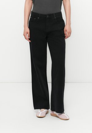 Persona che indossa jeans neri a gamba dritta, un top scuro e ballerine rosa chiaro con dettagli bianchi, in piedi su uno sfondo bianco.