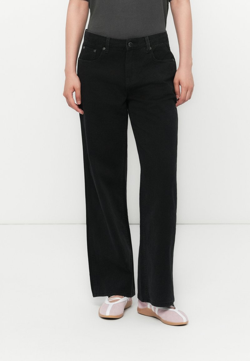 Person iført sorte straight-leg jeans, en mørk top og lyserøde balletflade med hvide detaljer, der står foran en hvid baggrund.