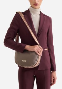 Borsa a tracolla grigia in nylon con tracolla rosa con motivo e logo dorato, caratterizzata da una forma arrotondata e una texture morbida, tenuta da una persona.