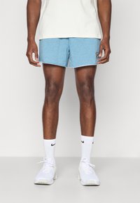 Gaiši zili sporta šorti ar teksturētu audumu, saplēstu apakšmalu un logo uzšuvumu. Apvienoti ar baltiem sporta apaviem un kokvilnas ceļgala zeķēm.