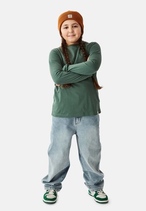 Grünes langärmliges Hemd, große hellblaue Jeans, braune Strickmütze und grün-weiße Sneakers. Geflochtene Haare sichtbar.