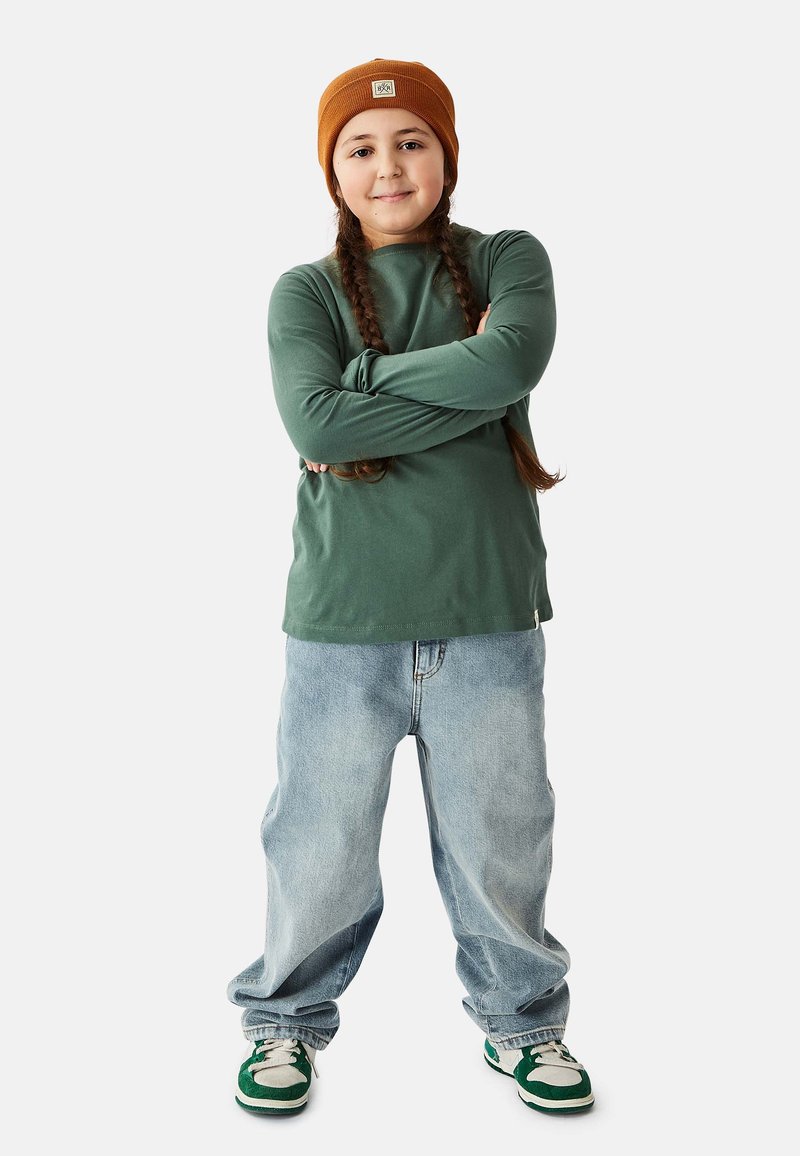 Green long-sleeve shirt, large light blue jeans, brown knit beanie, and green and white sneakers. Braided hair visible.