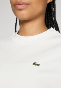 Kvinne med flettet hår iført en hvit skjorte med en liten grønn krokodille-logo på brystet.