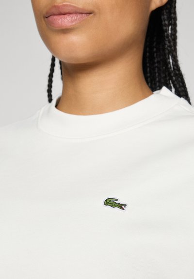 Femme avec des cheveux tressés portant une chemise blanche arborant un petit logo de crocodile vert sur la poitrine.