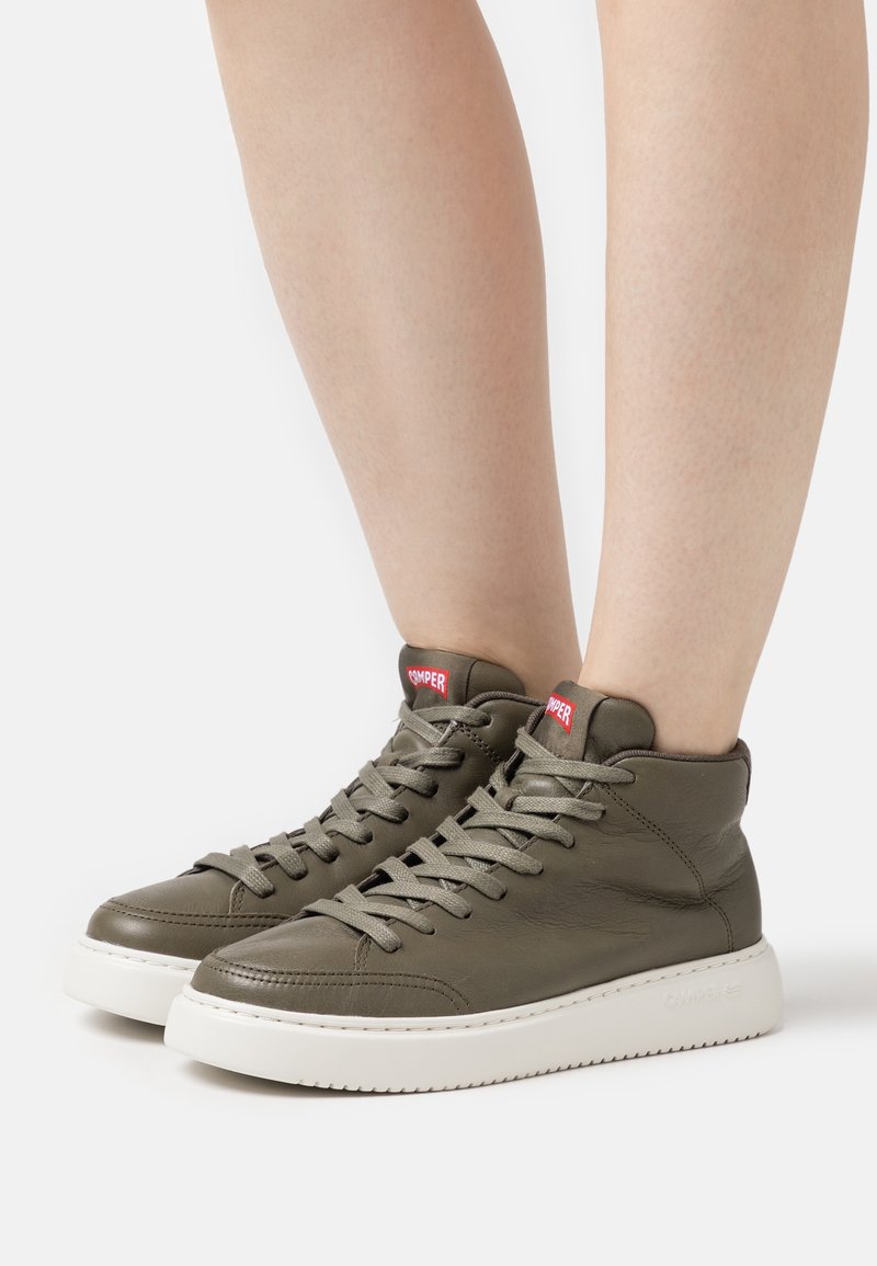 Personne portant des baskets montantes vert olive avec semelles blanches et logo rouge Camper sur la languette, debout sur un fond clair.