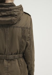 Giacca leggera verde oliva con cappuccio, vita regolabile e dettagli di cucitura a vista, dal fit rilassato e dalla texture morbida.