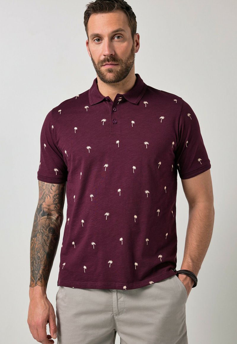 Mann mit Bart, der ein kastanienbraunes Poloshirt mit kleinen beigen Palmenmustern, beige Hose und einen Tattoo-Ärmel am linken Arm trägt.