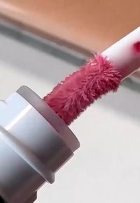 Applicateur de gloss à lèvres avec embout texturé et duveteux, recouvert d'un gloss scintillant mauve-rose, à l'intérieur d'un tube argenté.