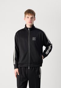 Zwarte zip-up trainingsjas met hoge kraag, witte zijstrepen en logo op de borst. Gemaakt van gladde materialen met een relaxte pasvorm.