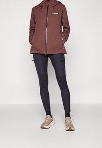 Personne portant une veste zippée bordeaux, des leggings noirs avec poches zippées, des chaussettes noires et des chaussures de sport beiges, debout devant un fond clair uni.