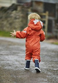 Combinaison de pluie orange imperméable avec détail de renard blanc sur la capuche, taille élastique et bottes imperméables marine. En extérieur, environnement humide.