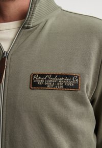 Grüne Zip-Jacke aus weichem Stoff, mit einem Aufnäher mit schwarzem und beige Text. Der Kragen hat eine gerippte Textur.