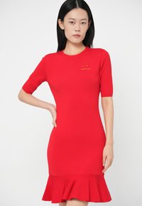 Γυναίκα στέκεται με το χέρι στη μέση, φορώντας εφαρμοστό κόκκινο φόρεμα κοντό μανίκι με φουσκωτό ποδόγυρο και μικρό κίτρινο λογότυπο στο στήθος.