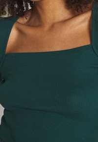 Ribbad grön topp med fyrkantig halsringning och figurnära design. Tyget verkar mjukt och texturerat, vilket framhäver plaggens form.