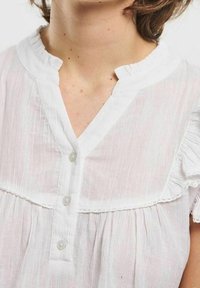 Donna che indossa una camicetta bianca leggera, a maniche corte, con colletto arricciato e tre bottoni davanti.
