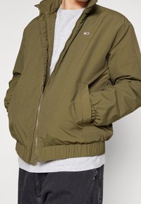 Giacca bomber verde oliva con chiusura a zip, polsini e vita elasticizzati. Presenta un piccolo logo sul petto e tasche laterali.