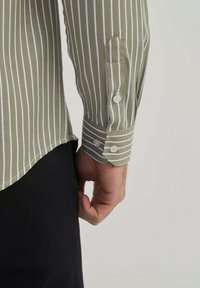 Chemise vert olive à rayures avec des rayures verticales blanches, poignets à boutons et coupe en bas de chemise, fabriquée en tissu texturé.