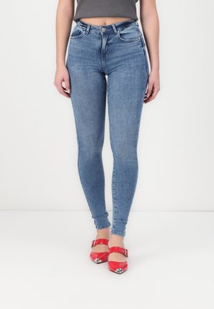 Femme porte un jean skinny bleu clair et des talons rouges à bouts pointus avec des brides à la cheville, debout sur un fond blanc.