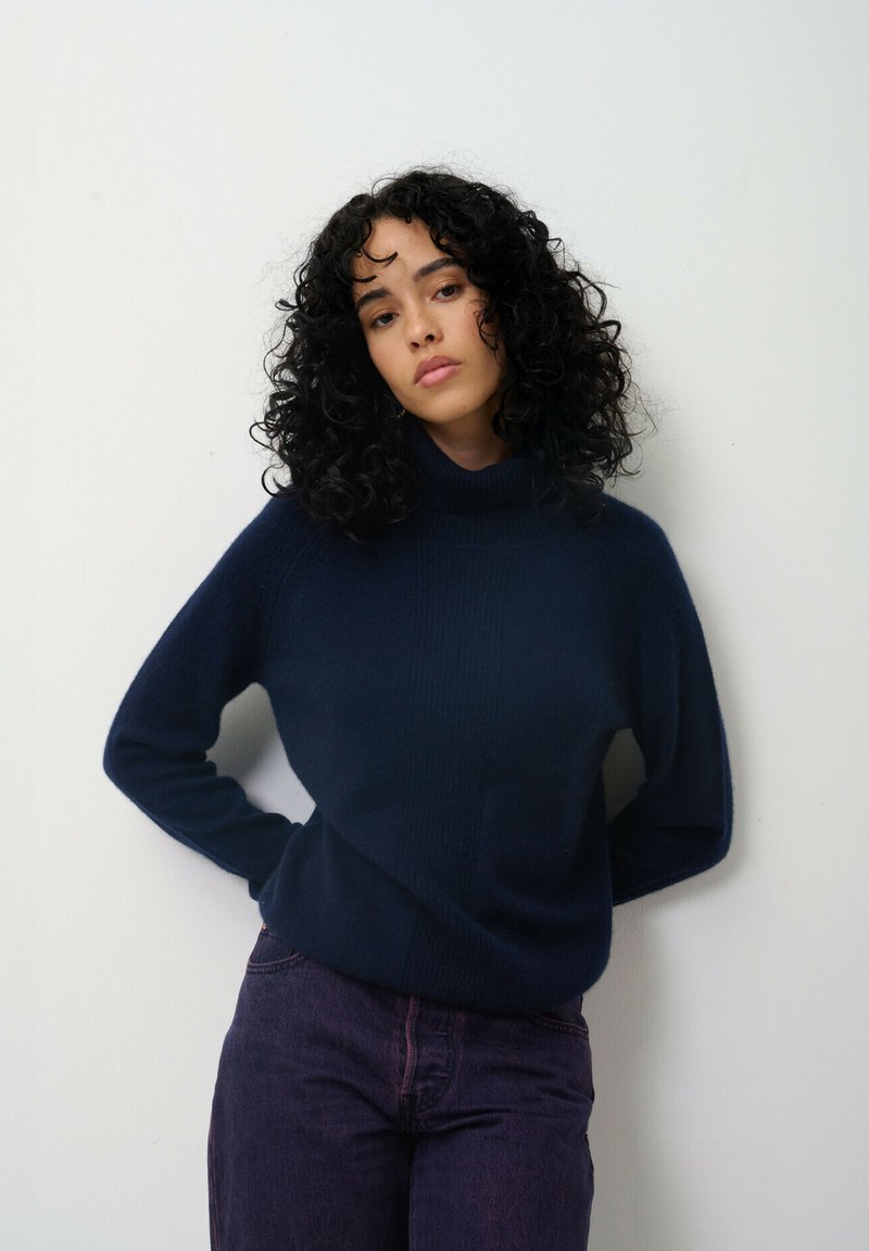 Marinblå tubhalsad tröja i mjukt material med ribbad textur och avslappnad passform. Kombination med mörklila jeans.