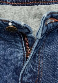Denim kavbojke z zadrgo, z sivo oblogo pasu. Temno modra tkanina, bakrena zadrga in vidni detajli v oranžni barvi.