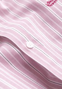 Tissu rayé rose et blanc avec un bouton blanc et un logo partiellement brodé en fil rouge sur le côté supérieur droit.