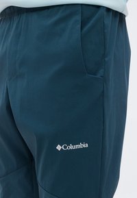 Σκούρο μπλε παντελόνια της μάρκας Columbia που φοριούνται από ένα άτομο, εμφανίζοντας την πλαϊνή τσέπη και το λογότυπο της μάρκας στο πάνω μέρος του μηρού.