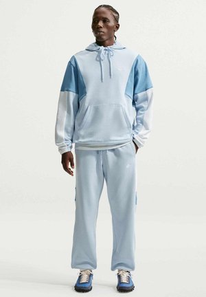 Man staat gekleed in een lichtblauwe en witte colorblock hoodie met bijpassende joggingbroek en blauwe sneakers, handen ontspannen langs het lichaam.