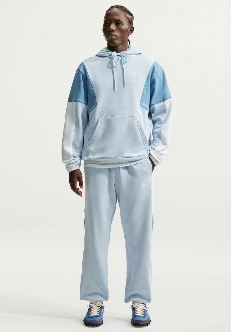 Man staat gekleed in een lichtblauwe en witte colorblock hoodie met bijpassende joggingbroek en blauwe sneakers, handen ontspannen langs het lichaam.