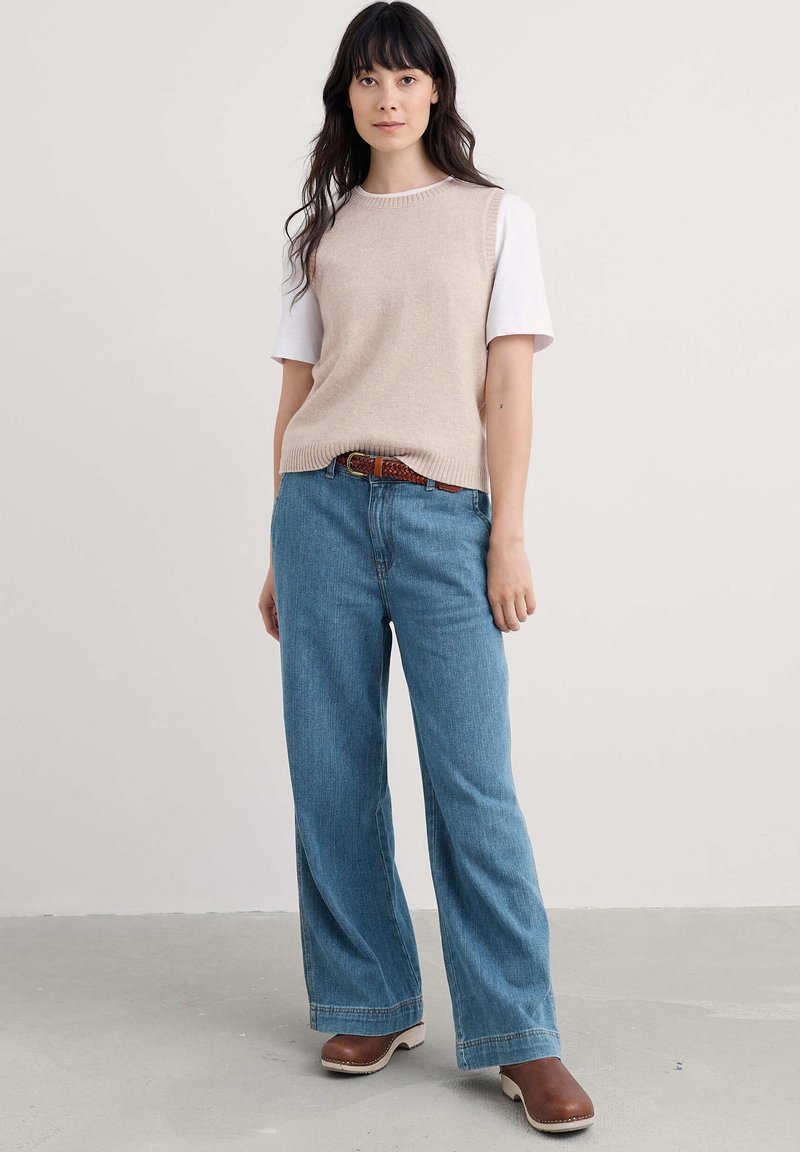 bézs kötött ujjatlan pulóver fehér póló felett, világoskék, bő szárú farmernadrággal és barna klumpákkal párosítva. Látható a textúrázott öv.