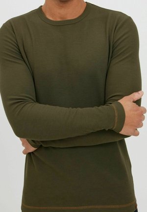 Hombre cruzando los brazos sobre el pecho, vistiendo una camiseta de manga larga acanalada color verde oliva con detalles de costura marrón en los puños y el dobladillo.