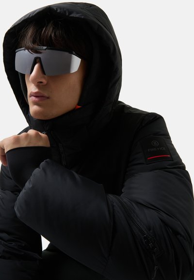 Black puffer jacket with a hood, featuring a zippered pocket, ribbed cuffs, and a logo patch. Model wears reflective sunglasses.