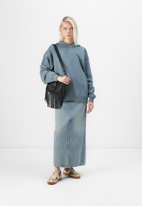 Sweat-shirt bleu à coupe décontractée, longue jupe en denim et baskets beiges. Sac à franges noir complète la tenue.