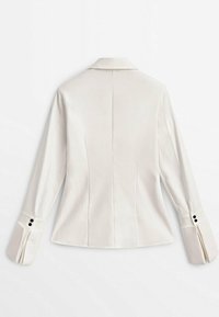 Veste blanche cintrée à manches longues, avec un détail de couture dans le dos, de larges poignets ornés de boutons noirs, et un col classique, présentée à plat vue de dos.