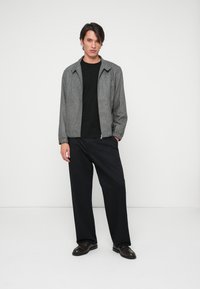 Grå, lynlåset jakke med en struktureret krave over en sort skjorte. Løse sorte bukser og mørke sko fuldender outfittet.