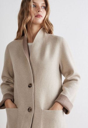 Jeune femme aux longs cheveux blonds portant un manteau en laine beige à deux boutons avec les mains dans les poches avant, regardant sur le côté.