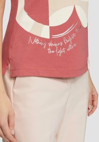 Röd bomullströja med abstrakta beiga former och vit text. Den nedre kanten har sidoflikar, i kombination med ljusrosa byxor.