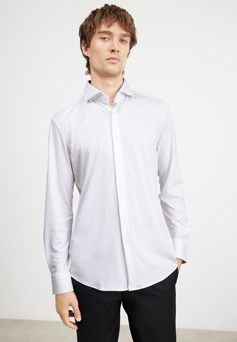 Maglietta bianca a maniche lunghe con colletto button-down, texture liscia e orlo arrotondato, abbinata a pantaloni neri.