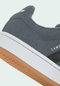 Zapatilla de ante gris con una entresuela blanca y una suela de goma marrón; presenta una franja diagonal negra y el texto "CAMPUS" en relieve.