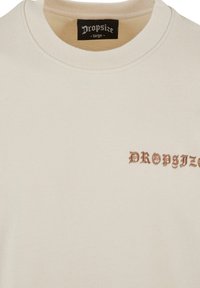 Lys beige sweatshirt med ribbet halsudskæring, der har "DROPSIZE" broderet i mørkebrunt på venstre side. Blødt bomuldsstof.