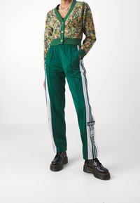 Vihreä kukkakuosiset neuletakki nappeilla, yhdistetty vihreisiin urheiluhousuihin, joissa on valkoiset sivuraidat ja Adidas-logo. Mustat massiiviset saappaat.