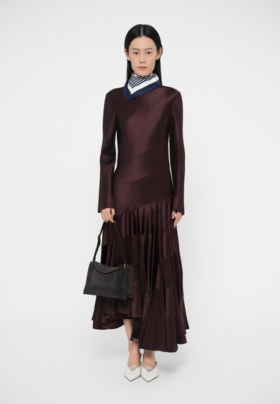 Bordeauxrode satijnen jurk met lange mouwen, asymmetrisch ontwerp en gelaagde ruches. Gestyled met een zwarte handtas en witte puntige schoenen.