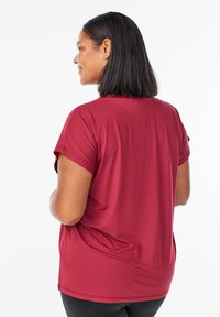 Žena s čiernymi vlasmi po pleciach, oblečená v červenom tričku s krátkym rukávom a čiernych nohaviciach, stojaca v profilu na bielom pozadí.