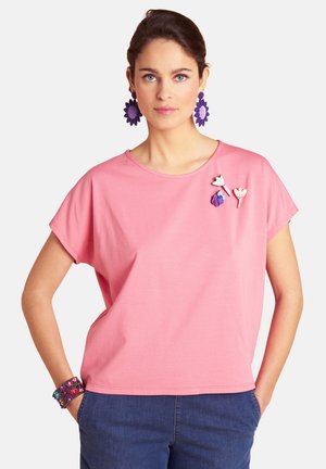 Frau trägt ein rosa kurzärmeliges Oberteil mit floralen Broschen, lila Blumenohrringen, Perlenarmband und blauen Hosen, steht mit Händen in den Taschen.