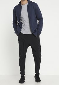 Felpa con zip colore navy sopra una maglietta grigia, abbinata a pantaloni neri a gamba affusolata e sneakers nere. Il tessuto sembra liscio con una vestibilità casual.