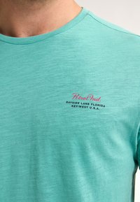 Turkoosi puuvillapaita, jossa on pyöreä kaula-aukko, ja jossa on pinkki ja musta teksti: "Petrol Ind. Bayside Lane Florida Key West Yhdysvallat."