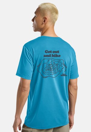 Mann mit kurzem blondem Haar, der ein blaues T-Shirt mit schwarzen Wanderkartenlinien und dem Text „Get out and hike“ auf der Rückseite trägt.