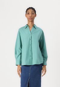 Camicia teal con colletto e maniche lunghe, abbinata a una gonna di jeans con spacco frontale. Tessuto dalla texture liscia.