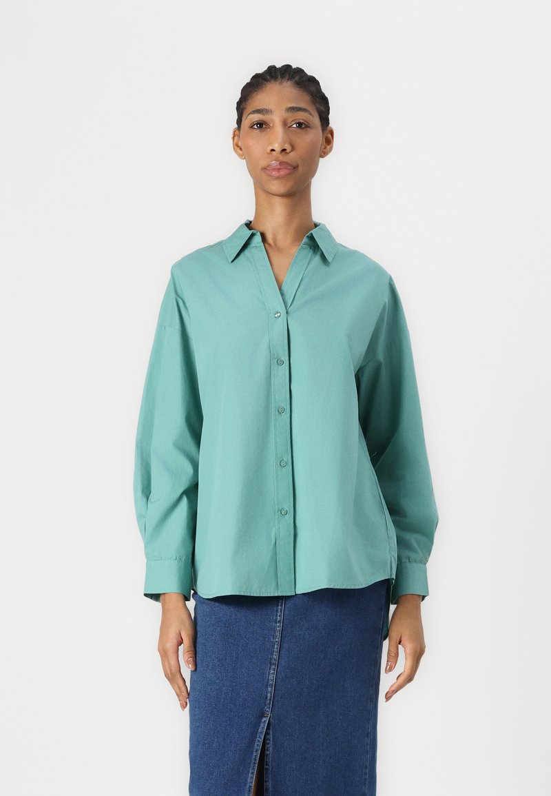 Camicia teal con colletto e maniche lunghe, abbinata a una gonna di jeans con spacco frontale. Tessuto dalla texture liscia.