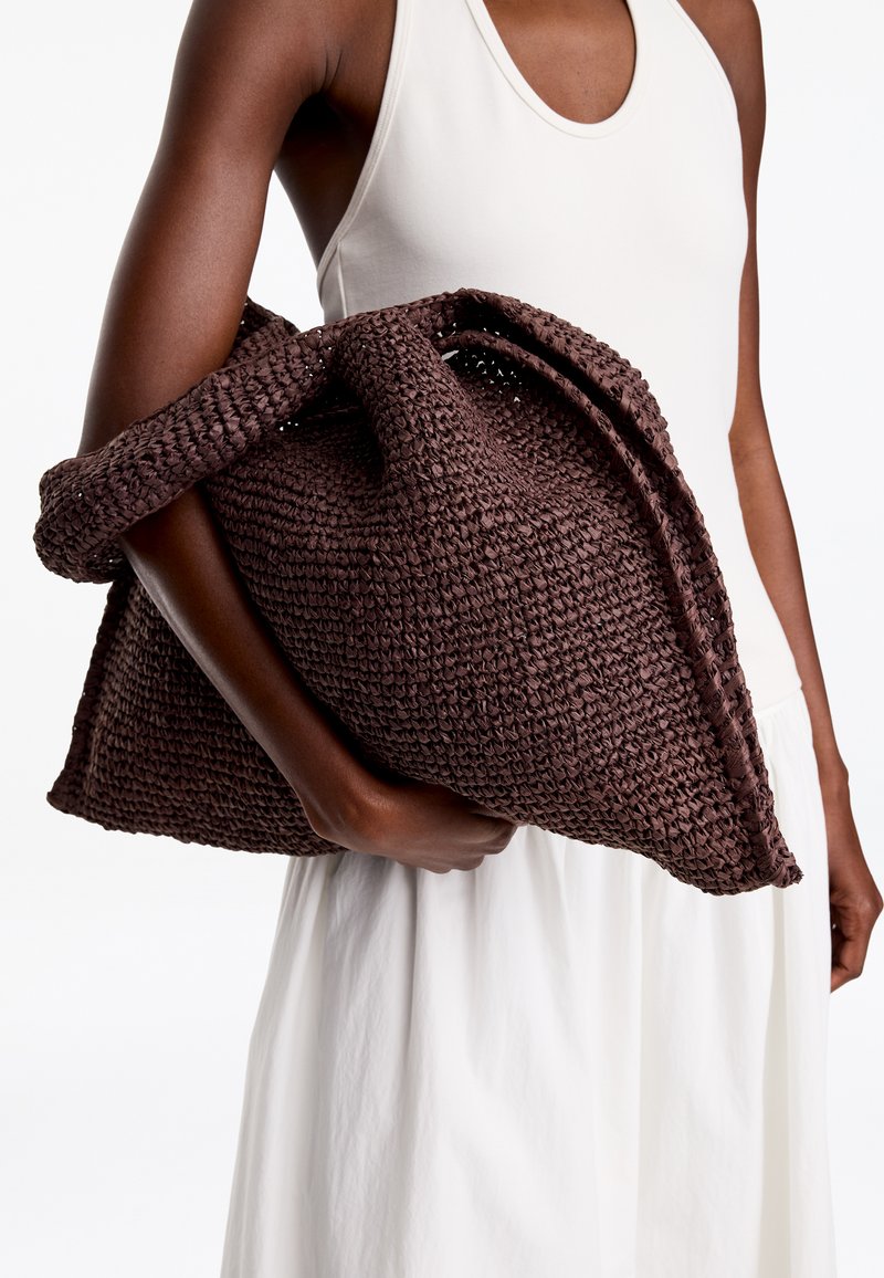 Personne en robe blanche sans manches tenant un grand sac tissé marron foncé sous le bras sur fond blanc.